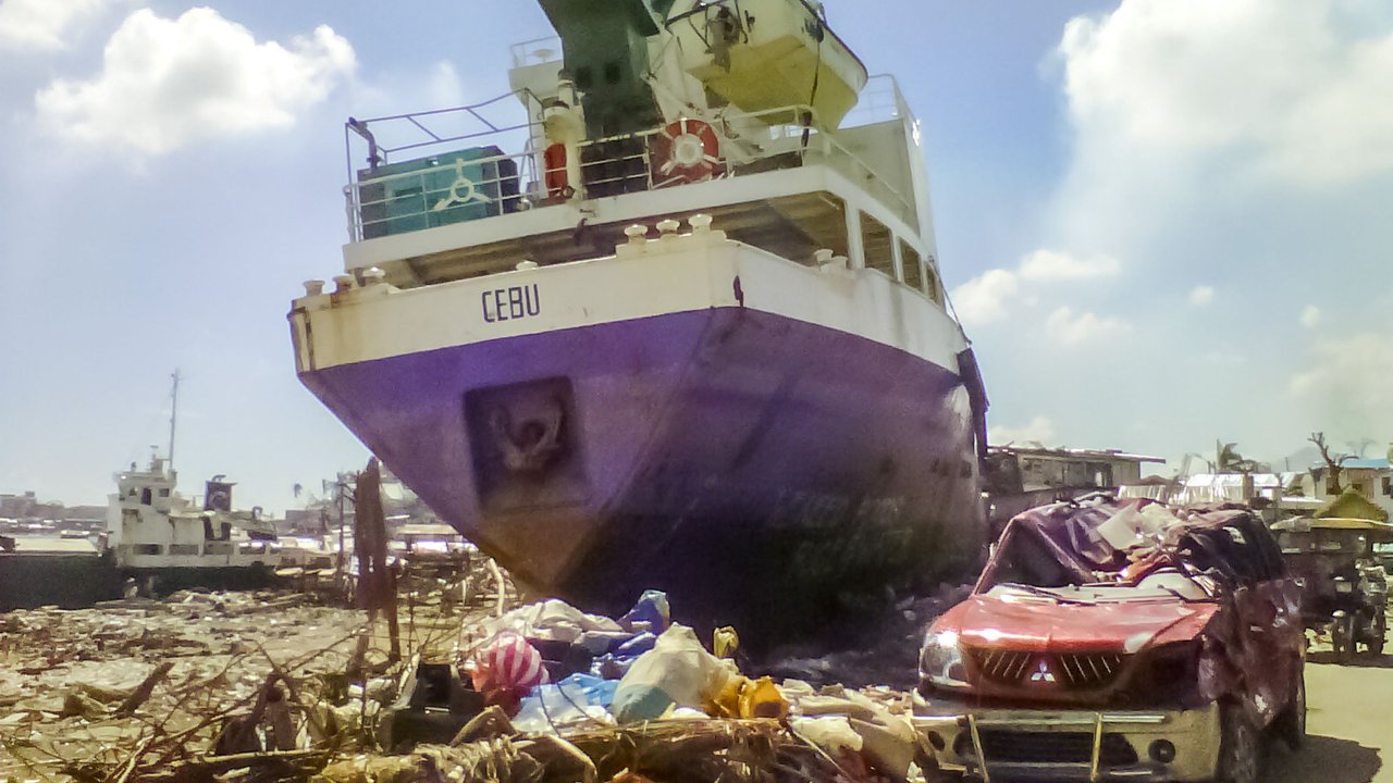 A large ship, labeled "CEBU," rests on a dock, partially surrounded by debris and trash. A damaged red vehicle is parked nearby, its body crumpled. The scene is illuminated by bright sunlight, with a clear blue sky and scattered clouds. The area reflects signs of neglect and environmental distress.
