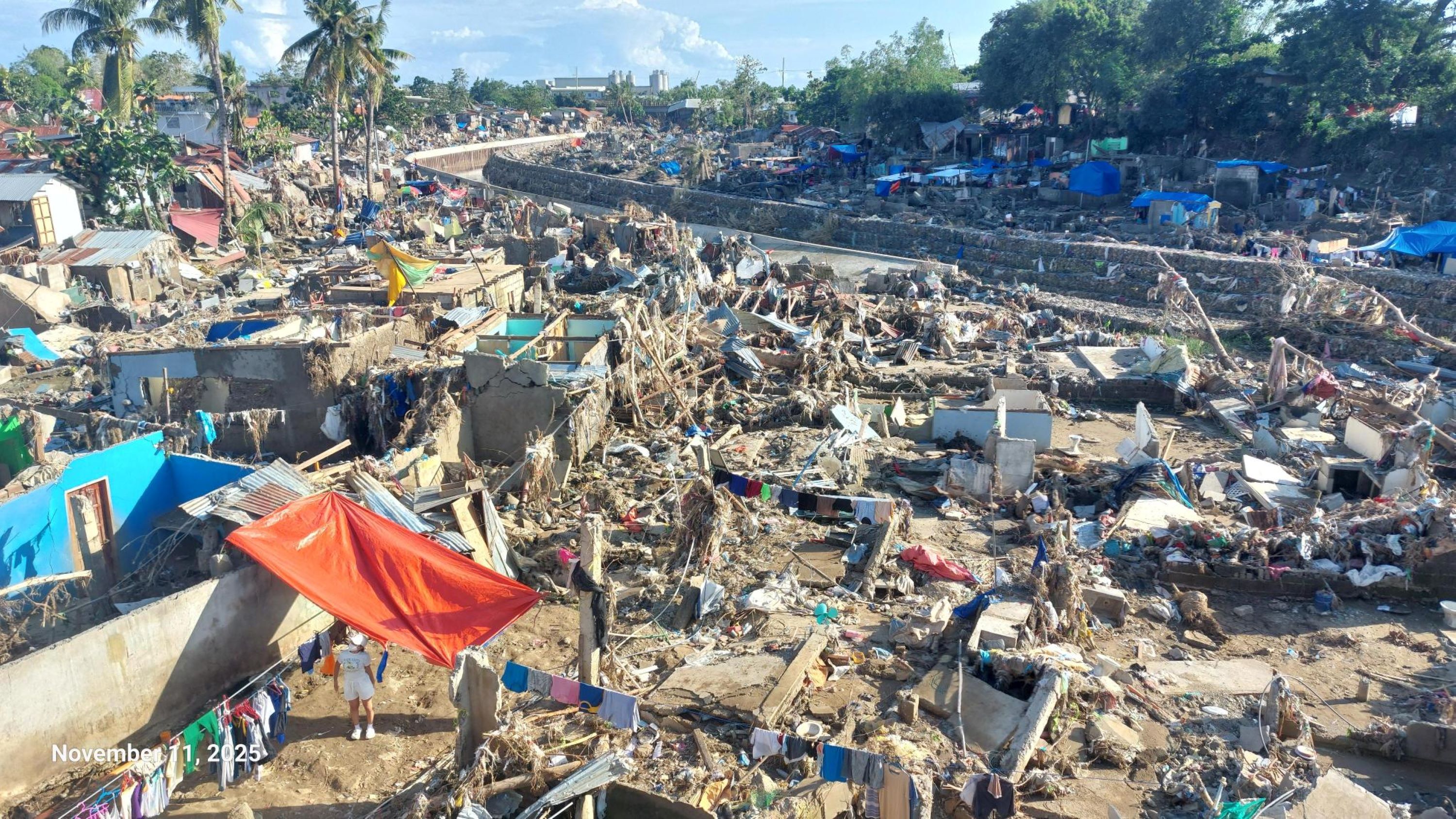 Aerial view of a devastated area shows numerous collapsed structures, debris, and remnants of homes. Brightly colored tarps and clothing hang on lines amidst the destruction. Palm trees are visible in the background, contrasting with the chaotic scene. The date in the corner indicates November 11, 2025.