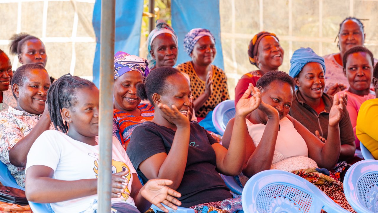 A group of women seated in chairs, some clapping, in a tented outdoor setting.