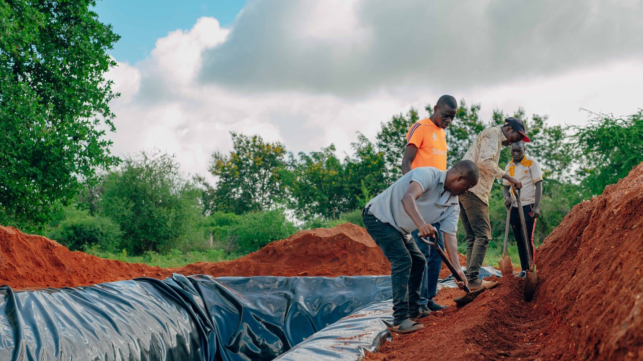 Three individuals are digging near a black plastic-covered trench in a green landscape under a cloudy sky.