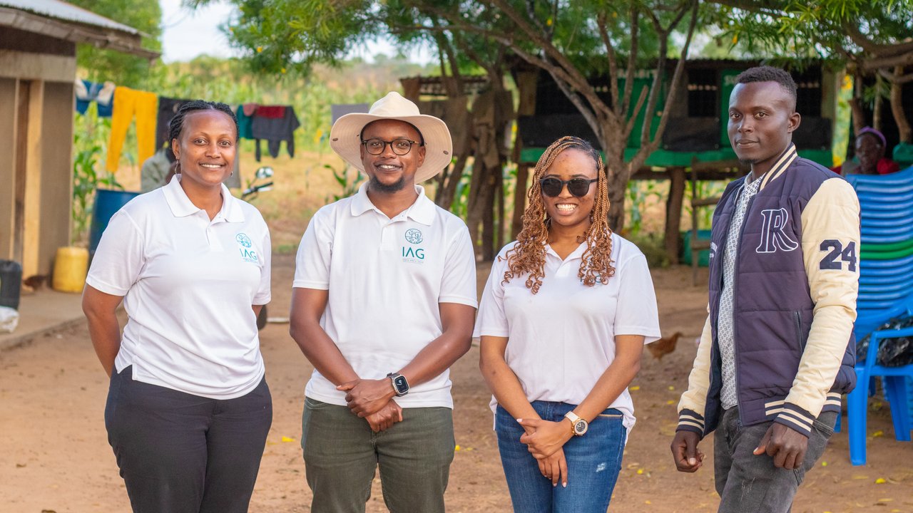 Four individuals in white shirts stand outdoors near a building, with laundry hanging in the background.