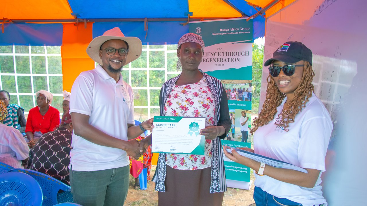 A man hands a certificate to a woman in a tent, with others seated in the background and banners displayed.