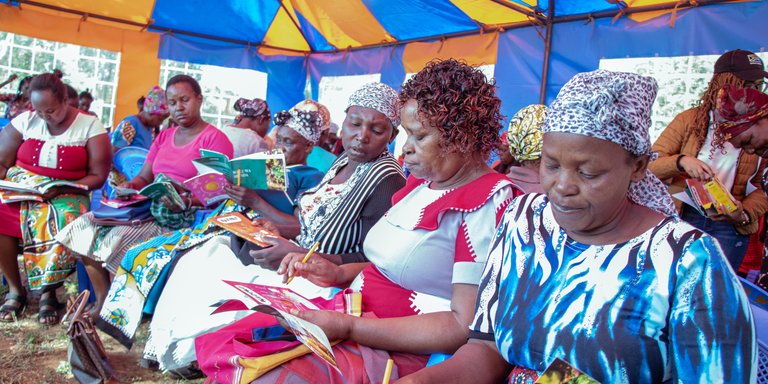 A group of women seated under a colorful tent, reading books and holding papers.