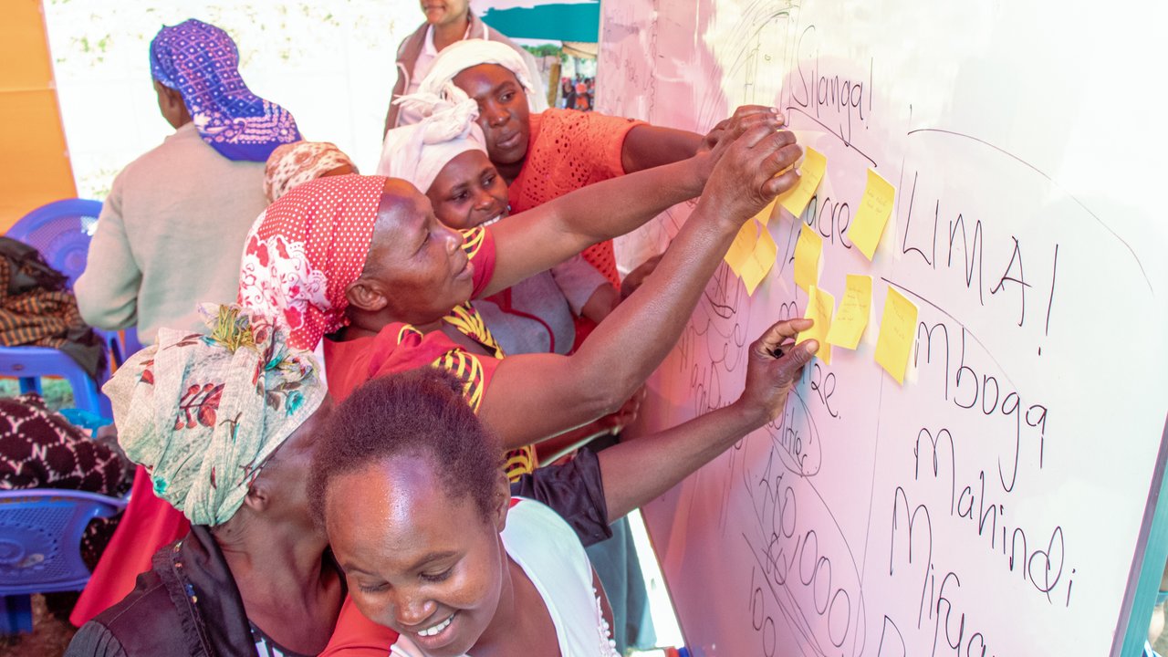 A group of people placing sticky notes on a whiteboard with handwritten text in a colorful indoor setting.
