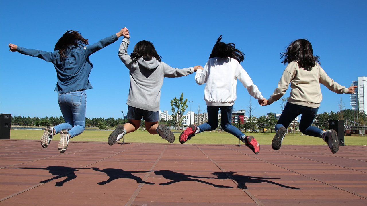 Group of friends jumping together outdoors Four people jump in unison, holding hands, against a clear blue sky and grassy field.