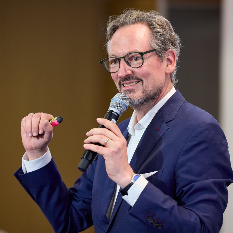 A speaker in a blue suit holds a microphone and a marker while addressing an audience.