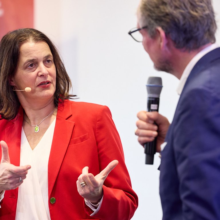 A woman in a red blazer gestures while speaking to a man holding a microphone in a professional setting.