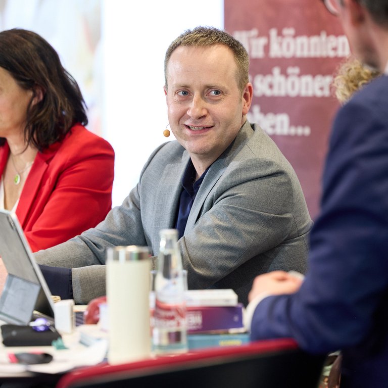 A panel discussion with three speakers at a table, surrounded by drinks and books, in a conference setting.