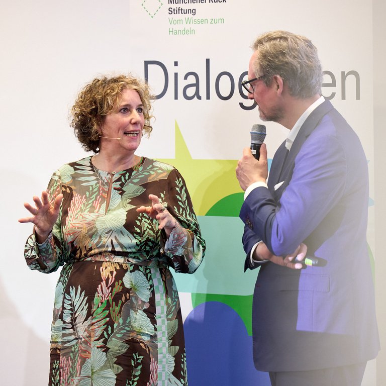 A woman gestures while speaking to a man holding a microphone at a dialogue event, with a branded backdrop.