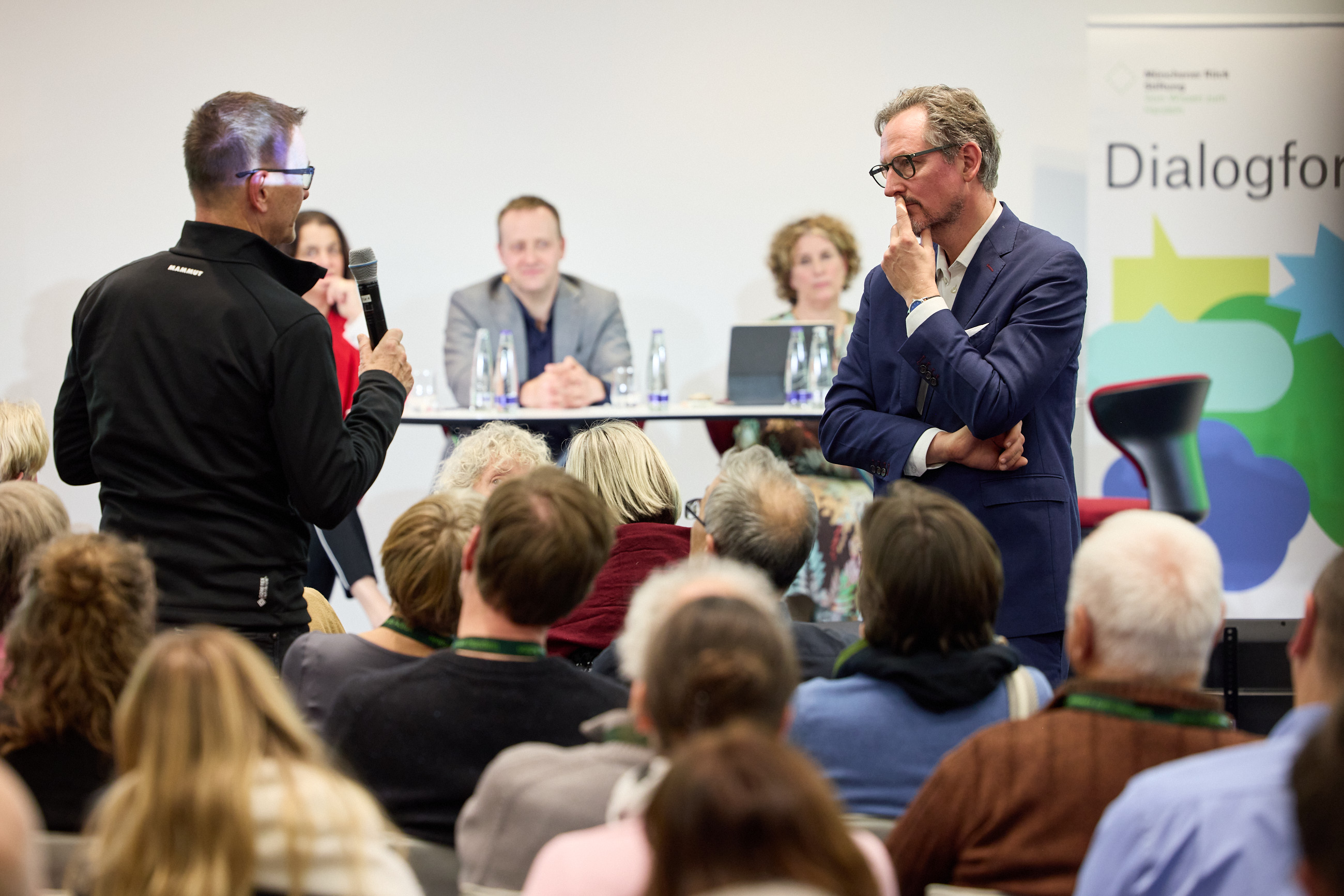 A speaker stands with a microphone addressing a panel of seated individuals during a discussion event.