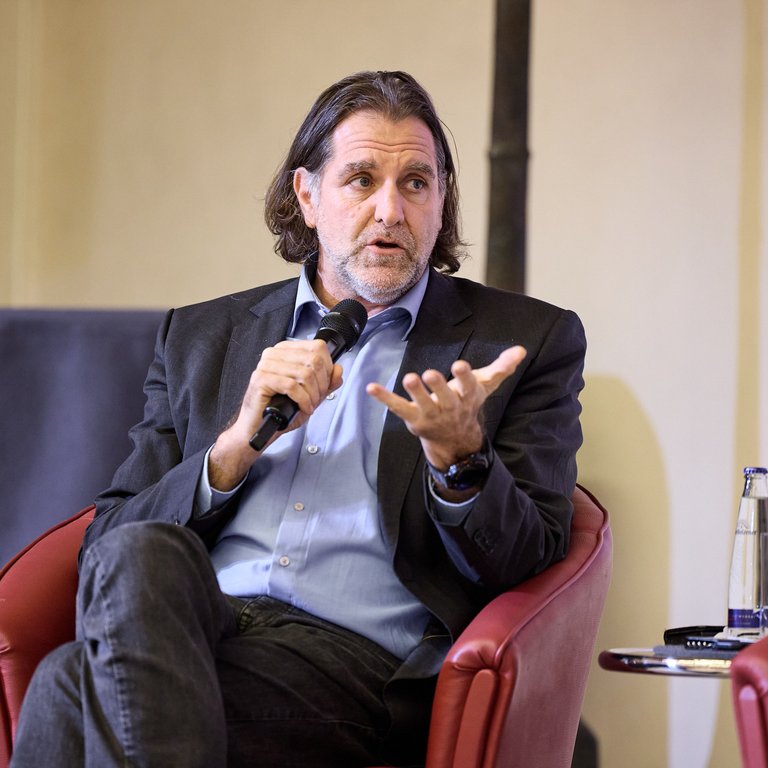 Ein Mann in einem Anzug sitzt in einem roten Sessel und spricht in ein Mikrofon, während er gestikuliert.