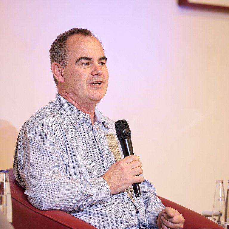 Ein Mann sitzt auf einem roten Sessel und hält ein Mikrofon in der Hand, im Hintergrund Wasserflaschen.