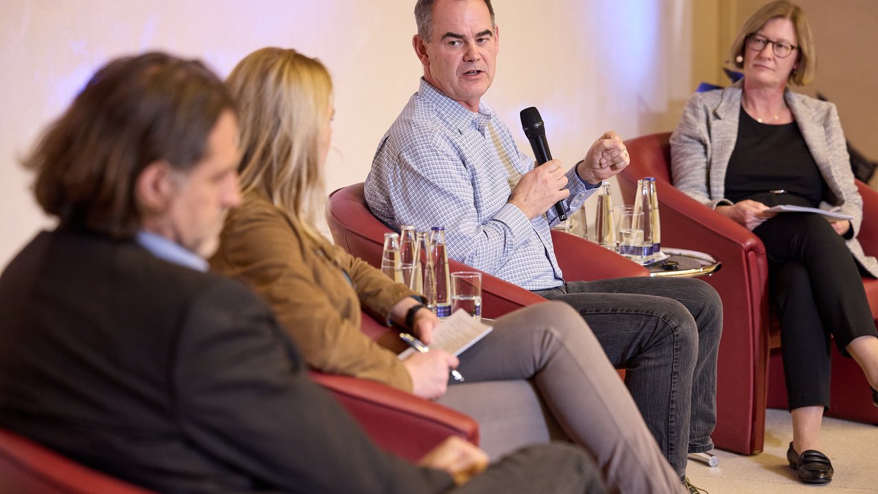 A panel discussion is taking place in a well-lit room with soft blue lighting. Participants are seated in modern red chairs, engaged in conversation. Water bottles and glasses are placed on a table in front of them. The atmosphere is professional, highlighting collaboration and dialogue among the speakers.