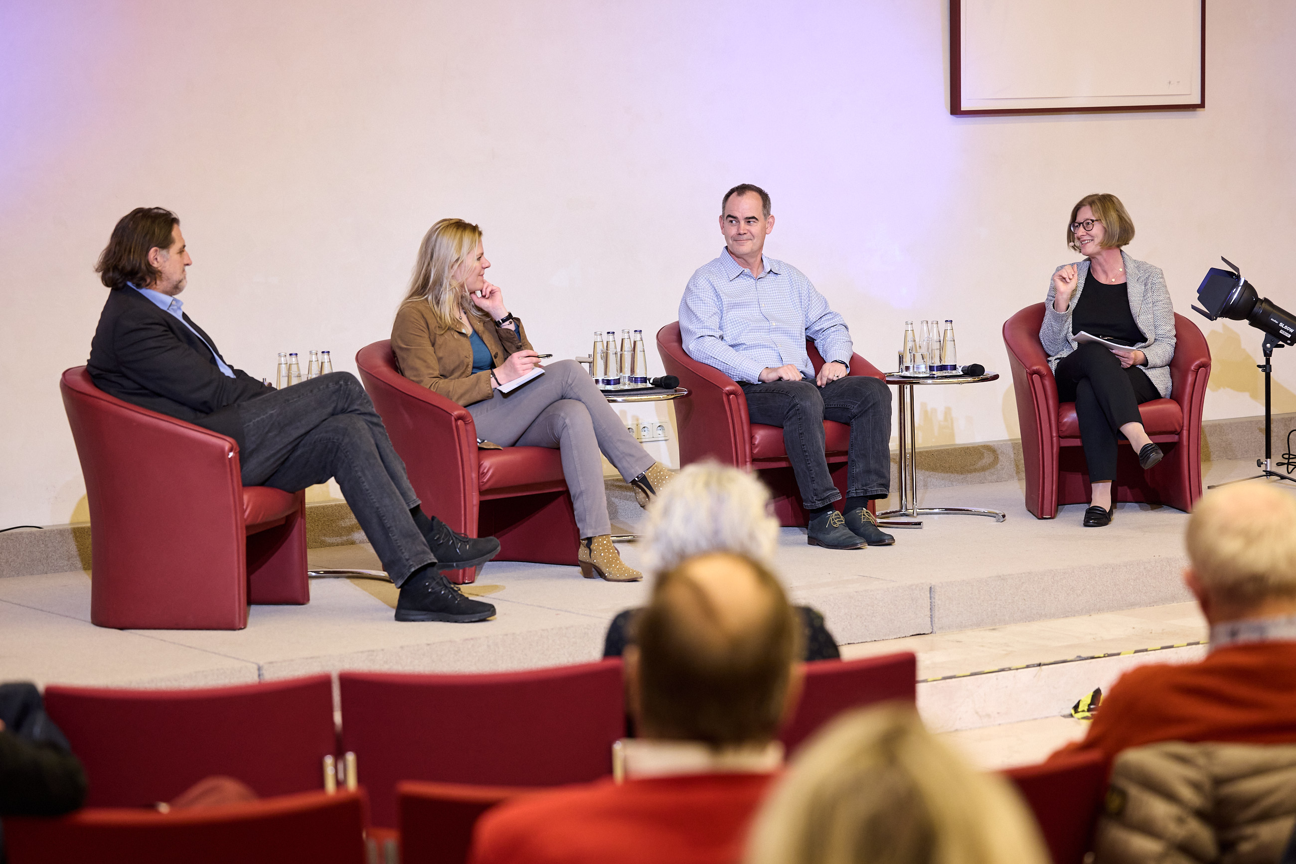 Vier Personen sitzen in roten Sesseln auf einer Bühne, mit Wasserflaschen und Notizen auf einem Tisch.