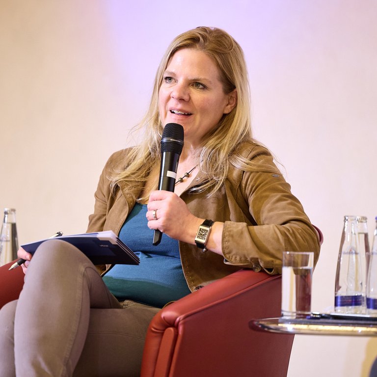 Eine Person sitzt auf einem roten Stuhl, hält ein Mikrofon und hat ein Notizbuch in der Hand. Wasserflaschen sind sichtbar.