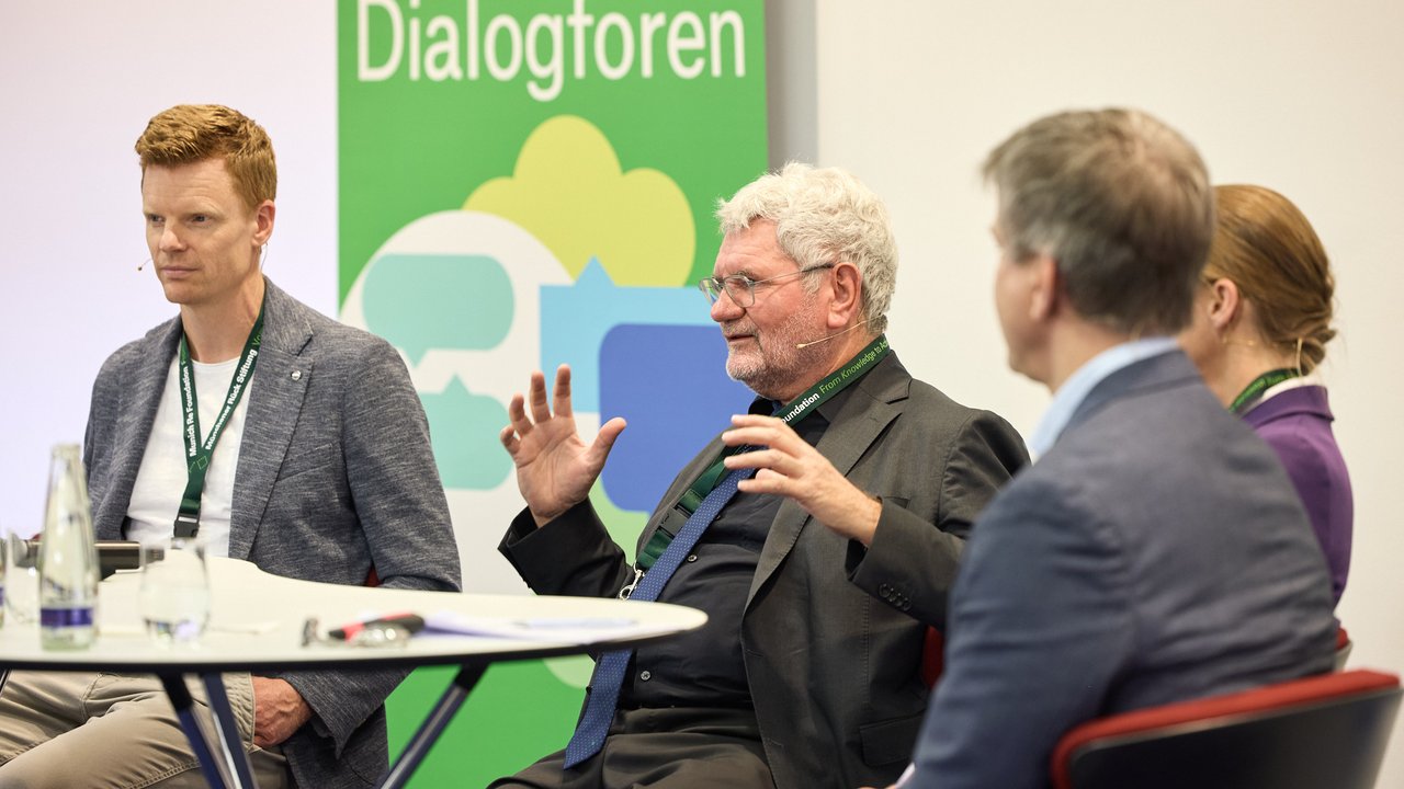 A panel discussion features four individuals seated around a circular table. The backdrop displays the word "Dialogforen" in bold green letters, accompanied by colorful abstract shapes. Participants are engaged in conversation, with one gesturing expressively. A glass water bottle and notebook are visible on the table.
