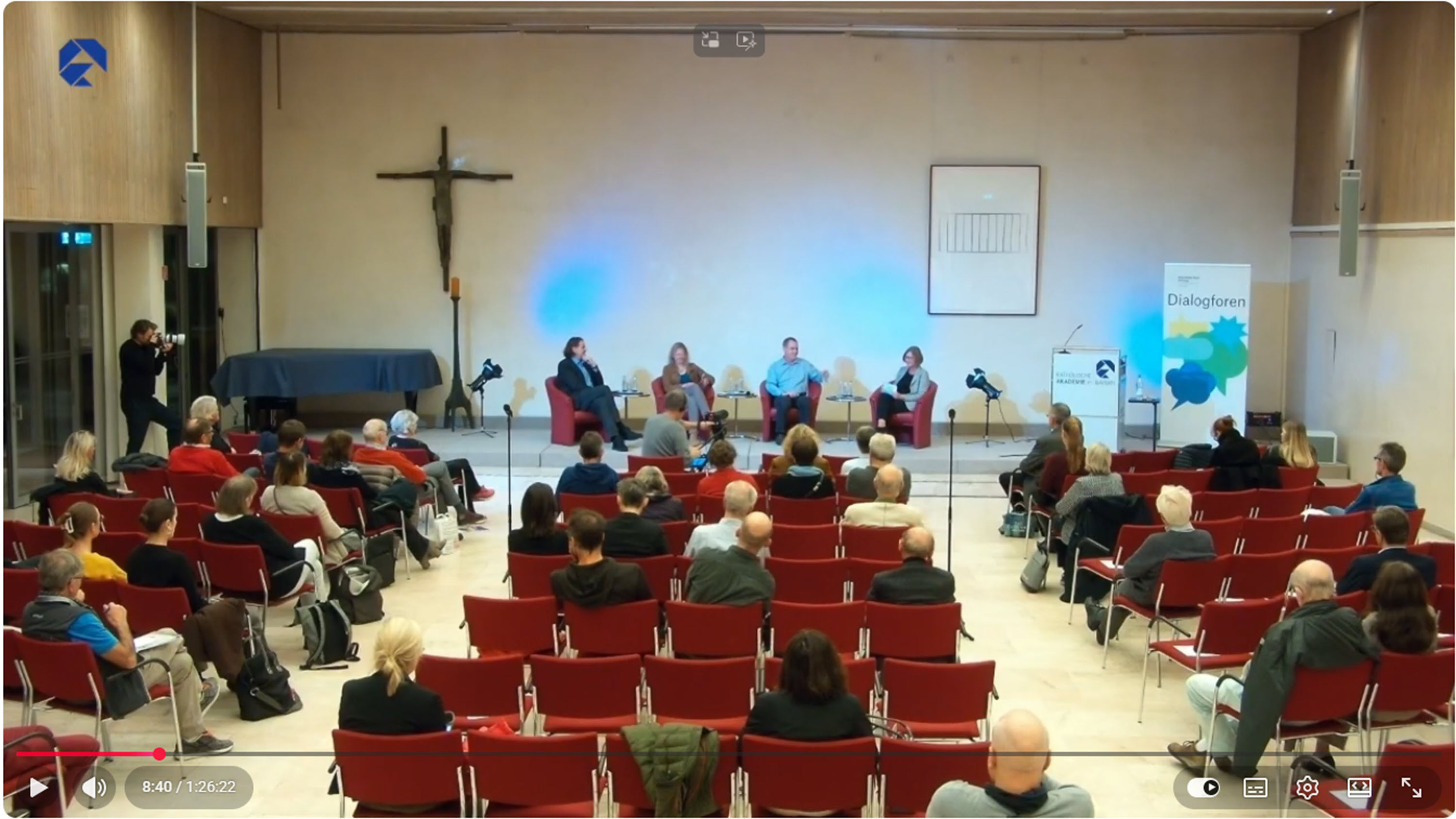 Eine Diskussionsrunde mit vier Personen auf roten Stühlen, Publikum sitzt in einem Raum mit Kreuz und Banner.