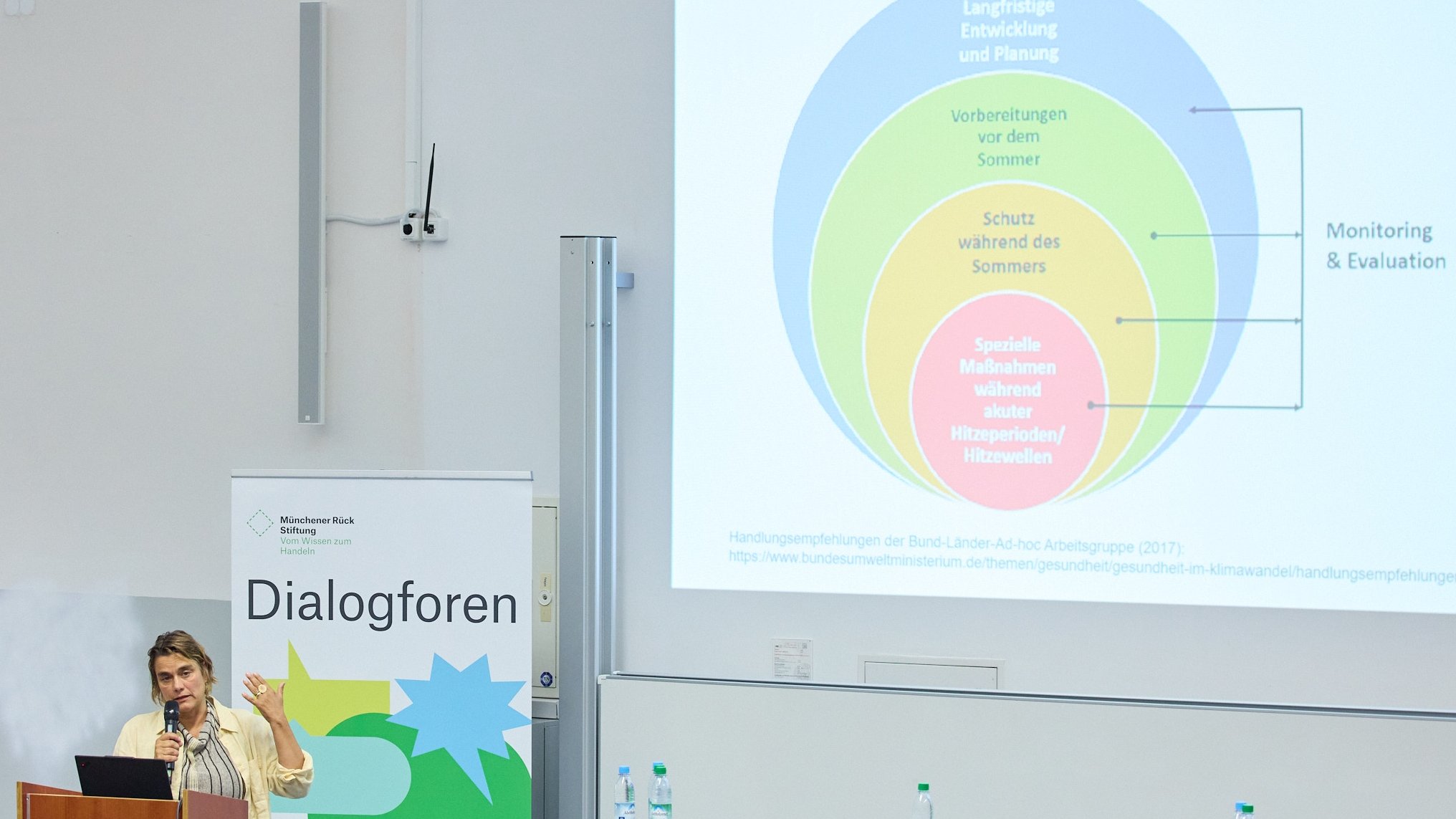 Eine Präsentation zu Hitzeschutzmaßnahmen in einem Seminarraum mit Zuhörern und einem Redner am Pult.