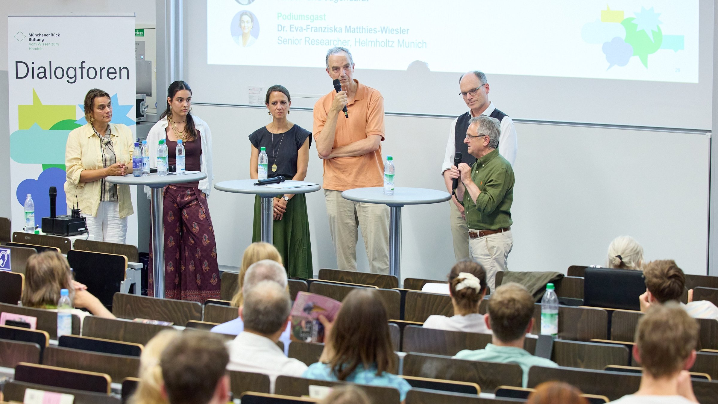 Eine Podiumsdiskussion mit fünf Sprechern in einem Hörsaal, Publikum sitzt in Reihen hinter ihnen.