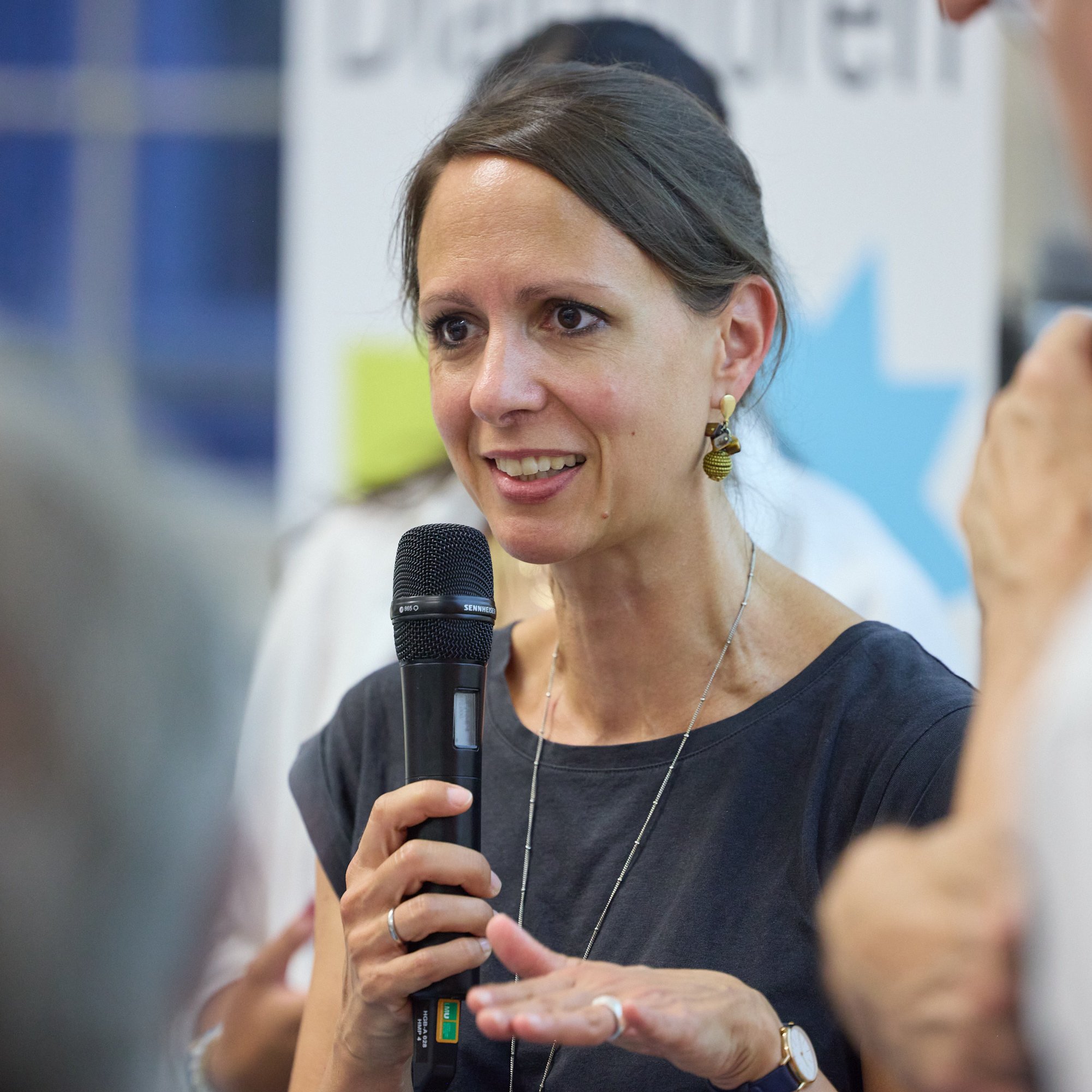 Eine Frau spricht mit einem Mikrofon in der Hand, im Hintergrund sind verschwommene Personen und ein Banner sichtbar.