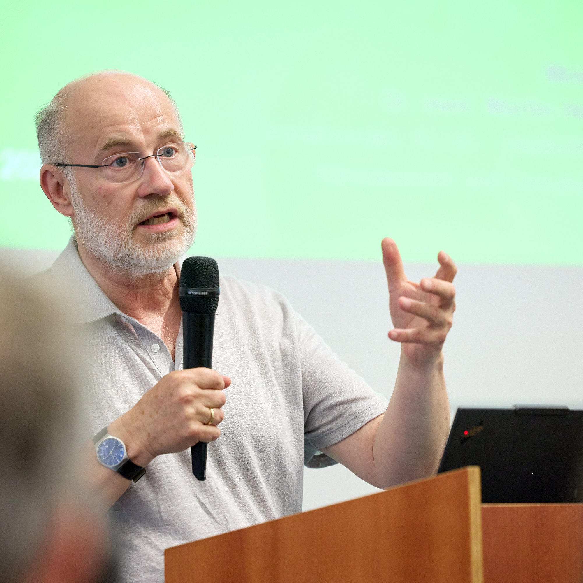 Ein Mann spricht mit einem Mikrofon, während er an einem Podium steht. Im Hintergrund ist eine grüne Präsentation sichtbar.