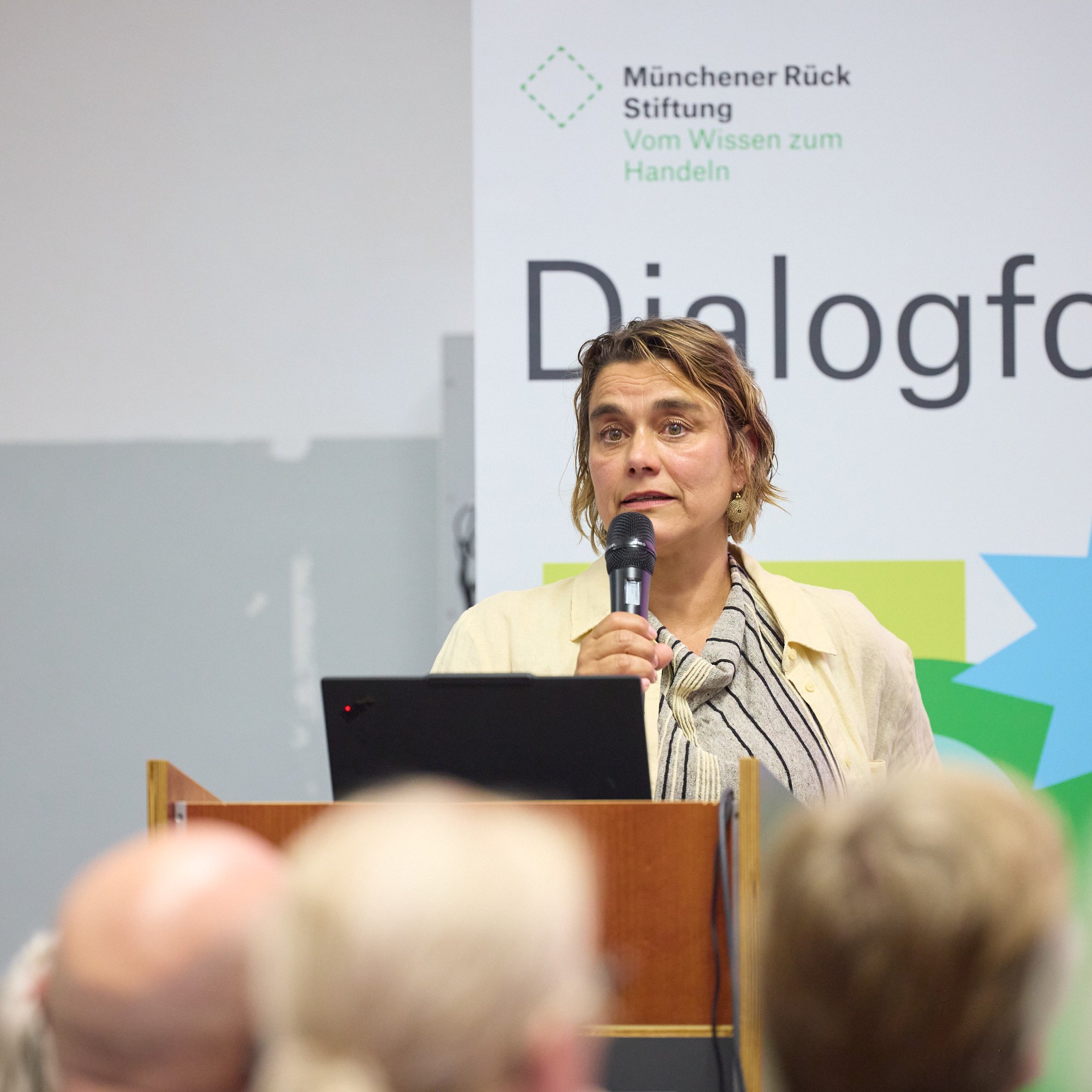Eine Frau spricht mit Mikrofon vor einem Podium, im Hintergrund ein Banner mit "Dialogforen".