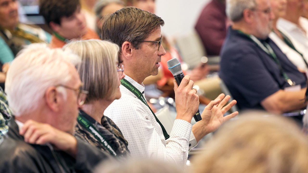 Eine Person hält ein Mikrofon und spricht, während andere Teilnehmer im Hintergrund zuhören.