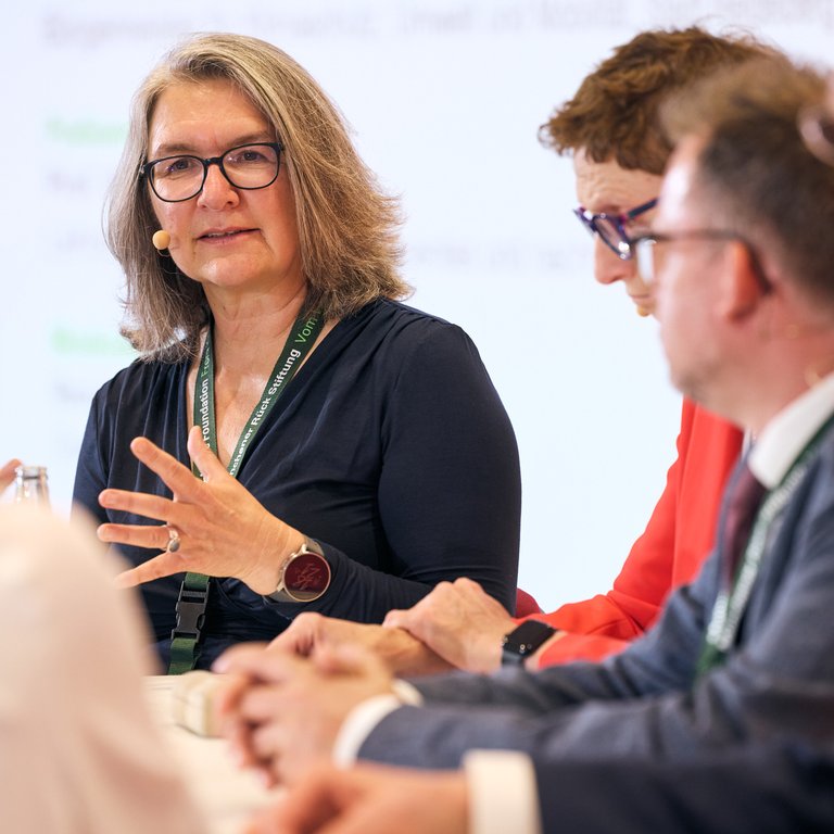Eine Frau spricht in einer Diskussionsrunde, umgeben von weiteren Personen an einem Tisch.