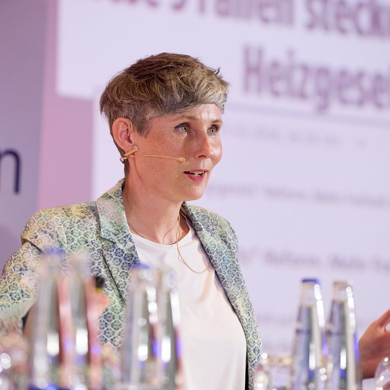 A speaker in a patterned blazer stands at a table with water bottles, presenting in front of a colorful backdrop.