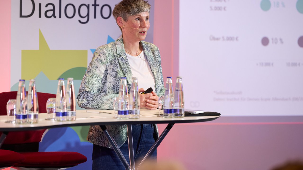 Eine Person steht an einem Tisch mit Wasserflaschen, im Hintergrund eine Präsentation mit Diagrammen.