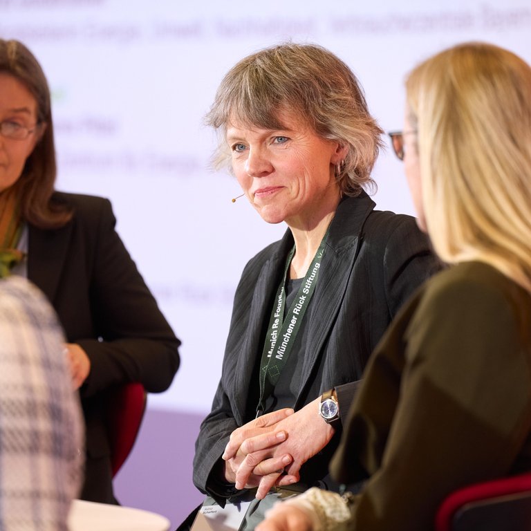 A group of individuals is engaged in discussion at a conference. One person, wearing a black blazer and a lanyard, sits with hands clasped, displaying a watch. Another individual, with long hair, listens attentively. The background features a colorful banner and a presentation screen, enhancing the professional atmosphere.