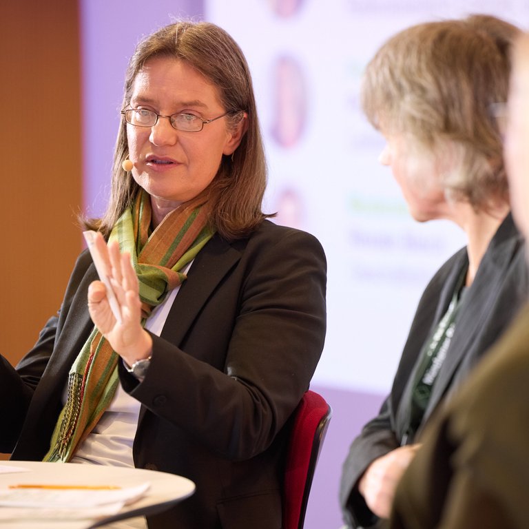 A woman gestures while speaking at a round table discussion, with two other women partially visible.