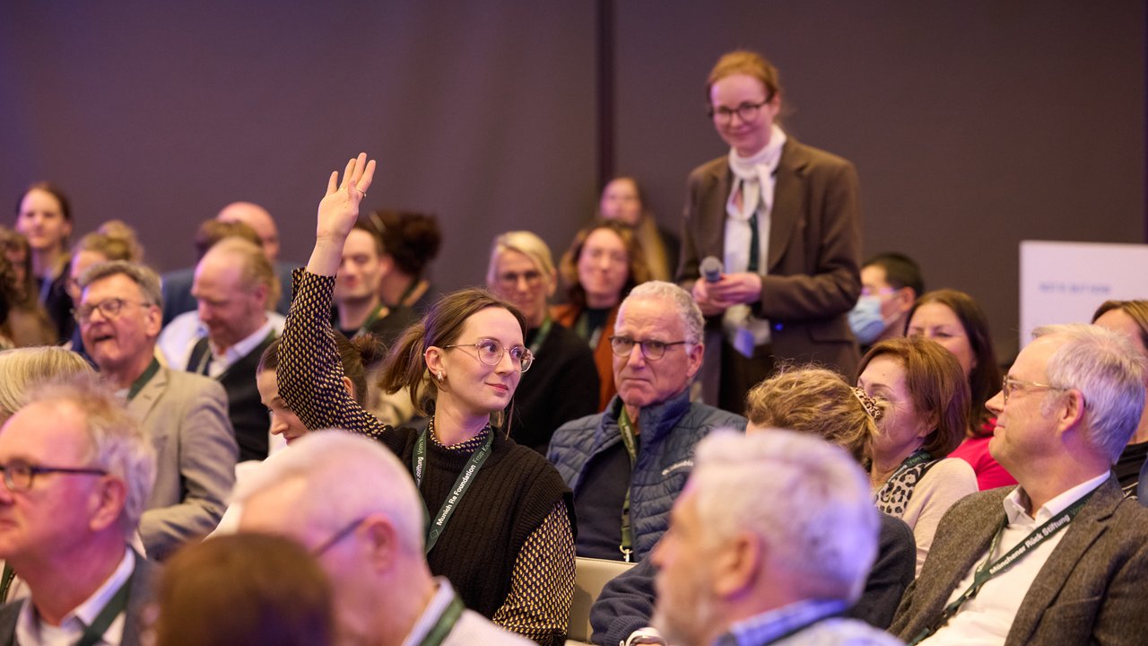 A conference audience with one person raising their hand, while another stands with a microphone.