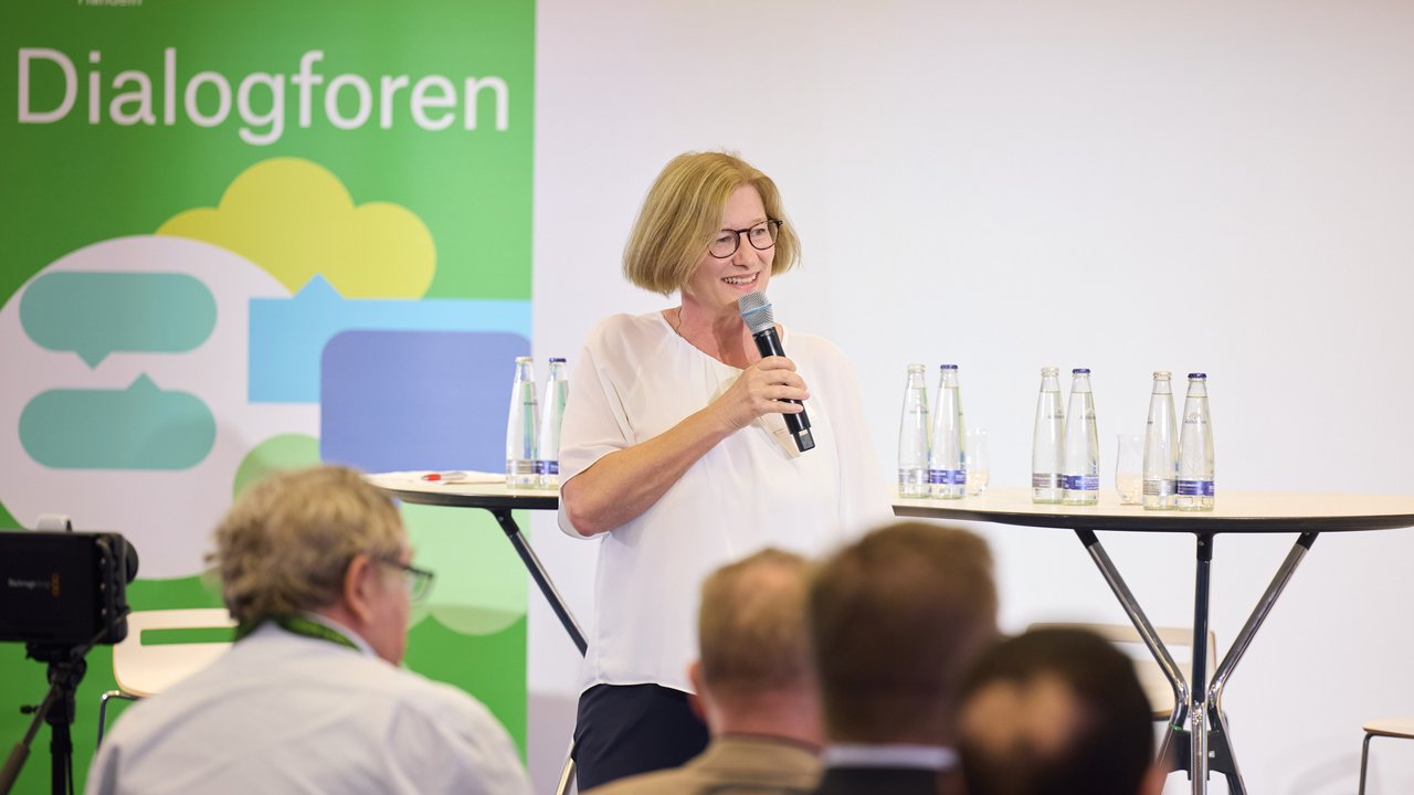 Eine Frau spricht mit Mikrofon vor einer Gruppe, auf einem Tisch stehen Wasserflaschen. Hintergrund mit "Dialogforen".