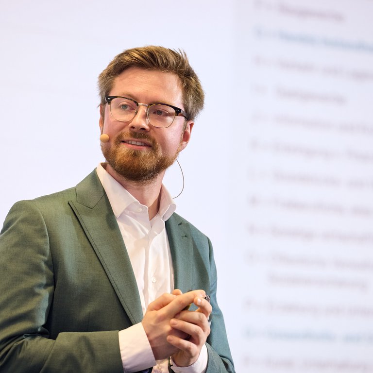 Ein Mann mit Brille in einem Anzug spricht vor einer Präsentation mit Diagrammen im Hintergrund.