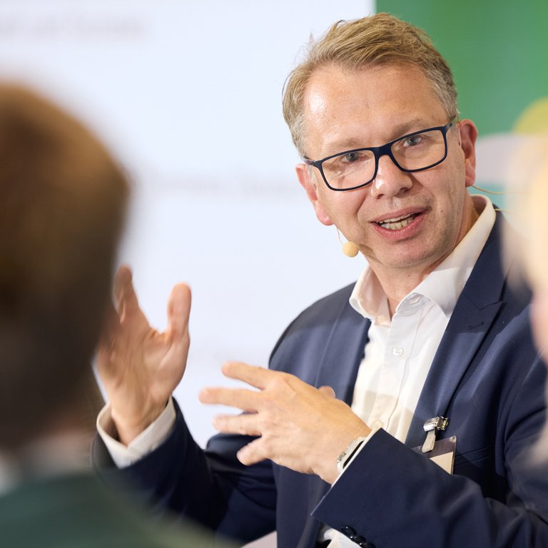 Ein Mann mit Brille spricht und gestikuliert, während andere Personen im Hintergrund unscharf zu sehen sind.