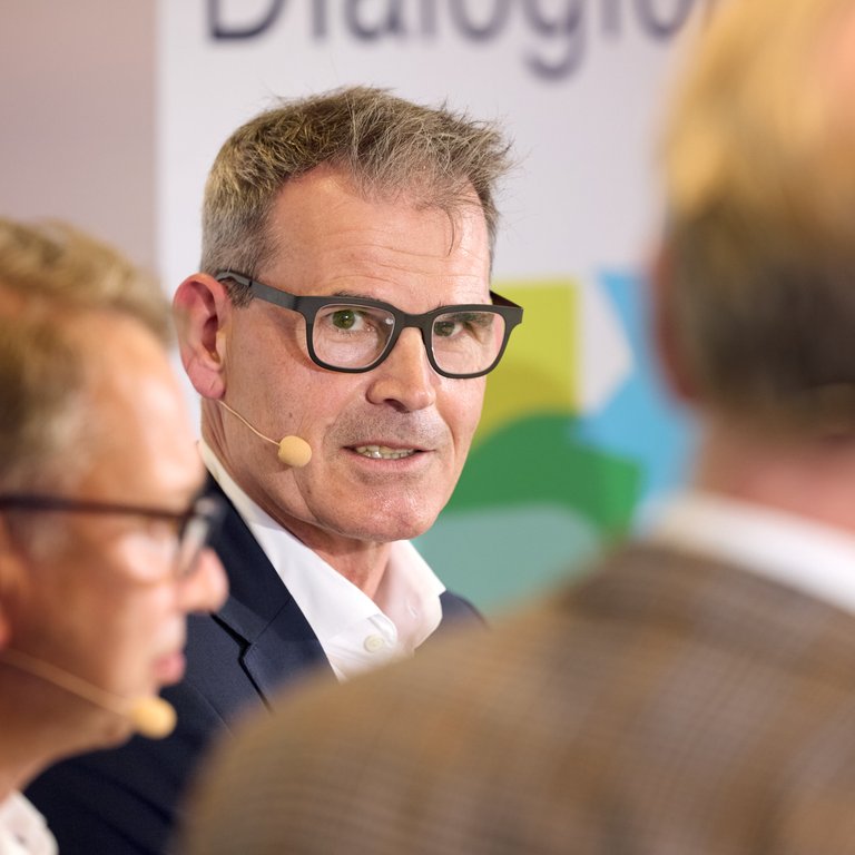 Ein Mann mit Brille und Mikrofon spricht, während zwei weitere Personen im Hintergrund sichtbar sind.