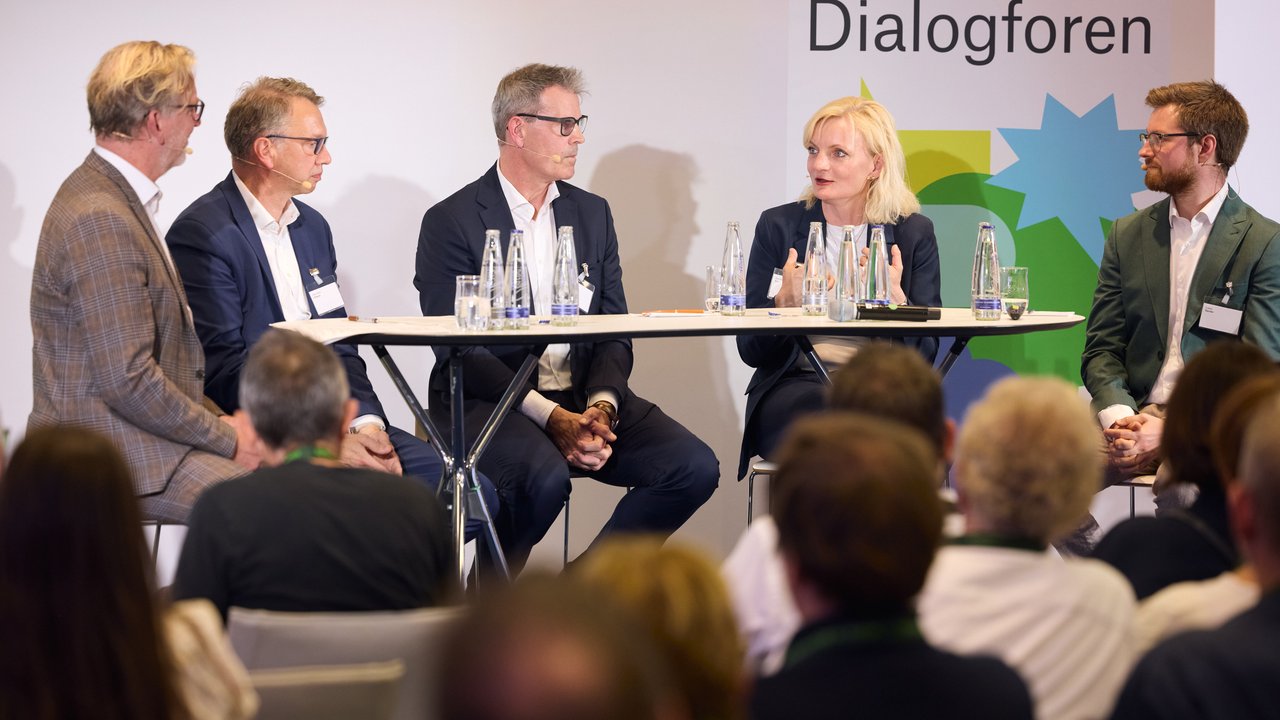 Vier Personen sitzen an einem Tisch während einer Diskussion, im Hintergrund ein Banner mit dem Wort "Dialogforen".