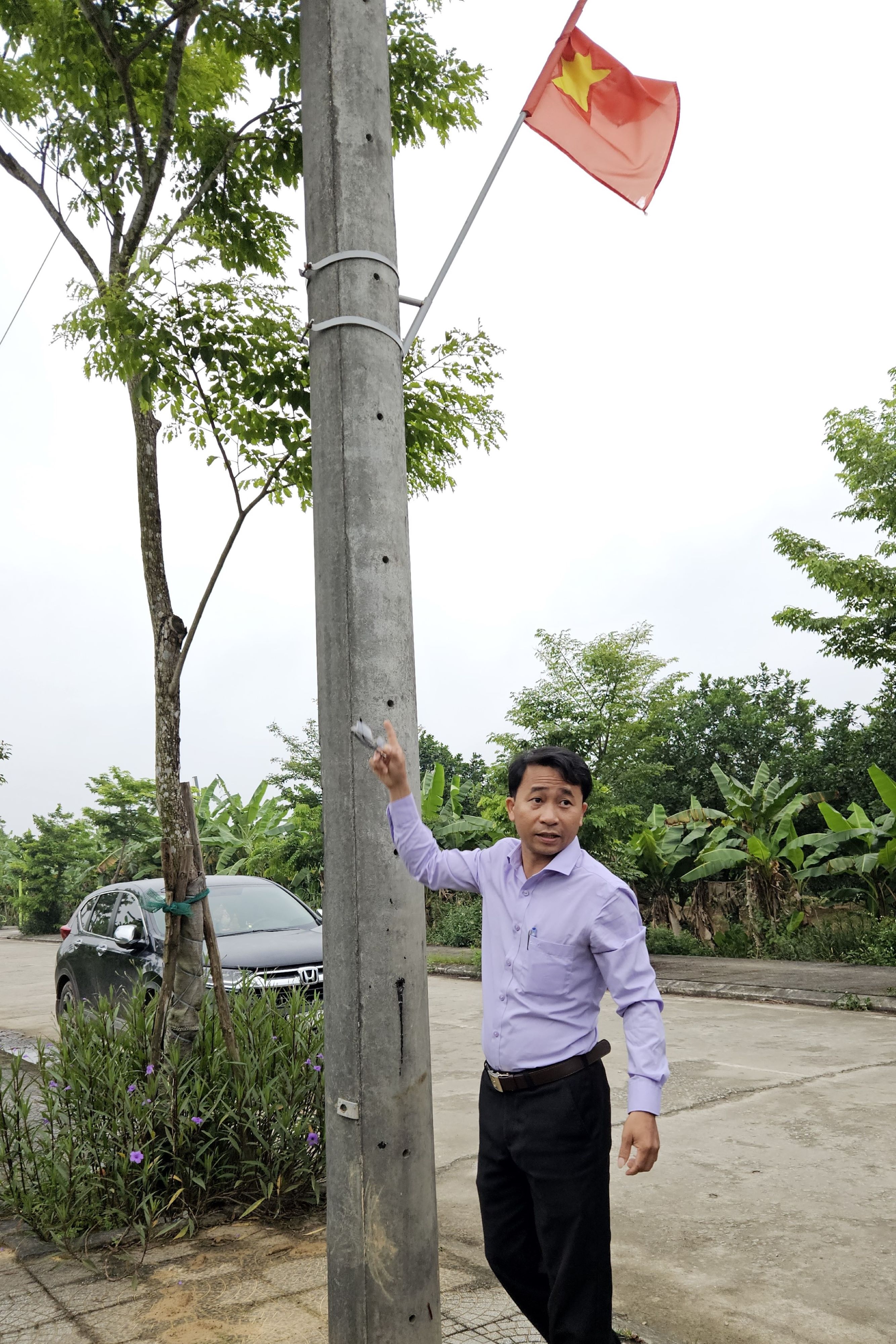 A person stands beside a concrete utility pole, gesturing towards a flag of Vietnam, which is attached to the pole. The background features lush greenery and a paved road, with a parked black car visible. The individual wears a light purple shirt and black pants, blending into the serene, natural setting.