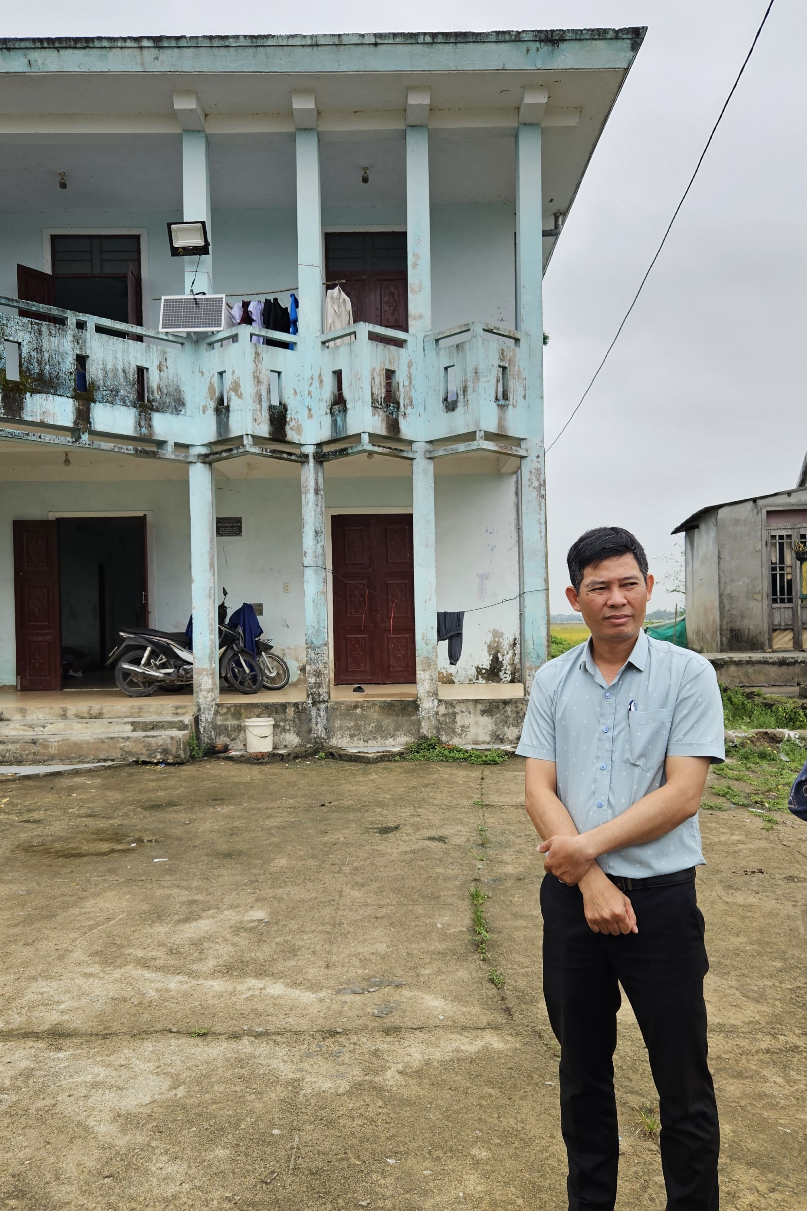 A weathered two-story building with peeling blue paint features multiple doors and a balcony. Clothes hang on a line above. In the foreground, a person stands with arms crossed, wearing a light blue shirt and dark pants. A motorcycle is parked nearby, and the ground is uneven with patches of grass.