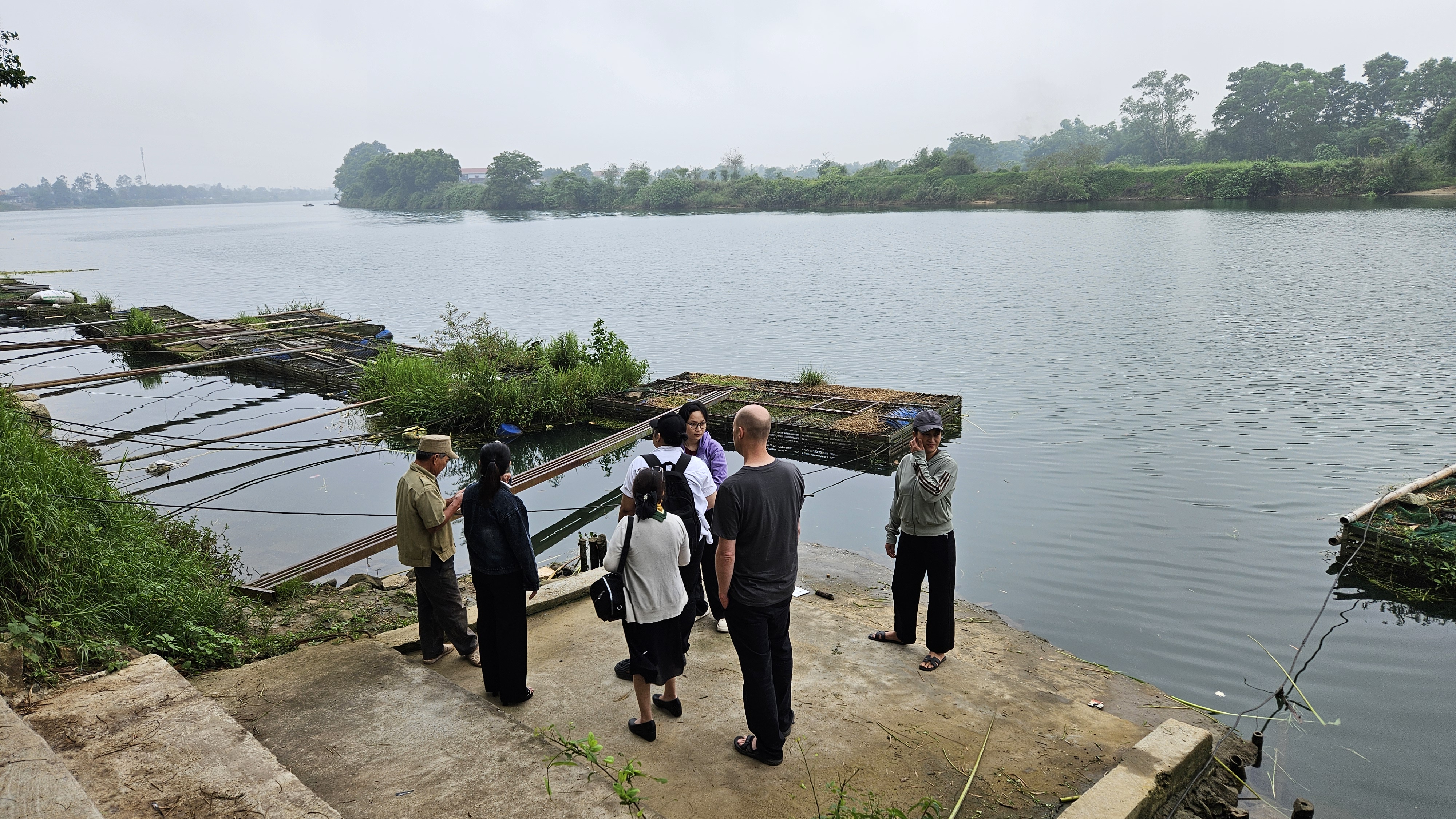 A group of individuals stands near a calm river, surrounded by lush greenery. Wooden cages with overgrown vegetation float on the water's surface. The atmosphere is tranquil, with a soft, cloudy sky above. The scene captures a moment of interaction among the group, emphasizing community and connection to nature.