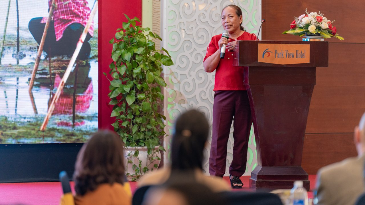 A speaker stands at a podium adorned with flowers, addressing an audience in a hotel setting. Behind them, a large screen displays a vibrant image of a person engaged in a water-related activity, surrounded by greenery. The room features decorative elements, including a patterned wall and potted plants, enhancing the atmosphere.