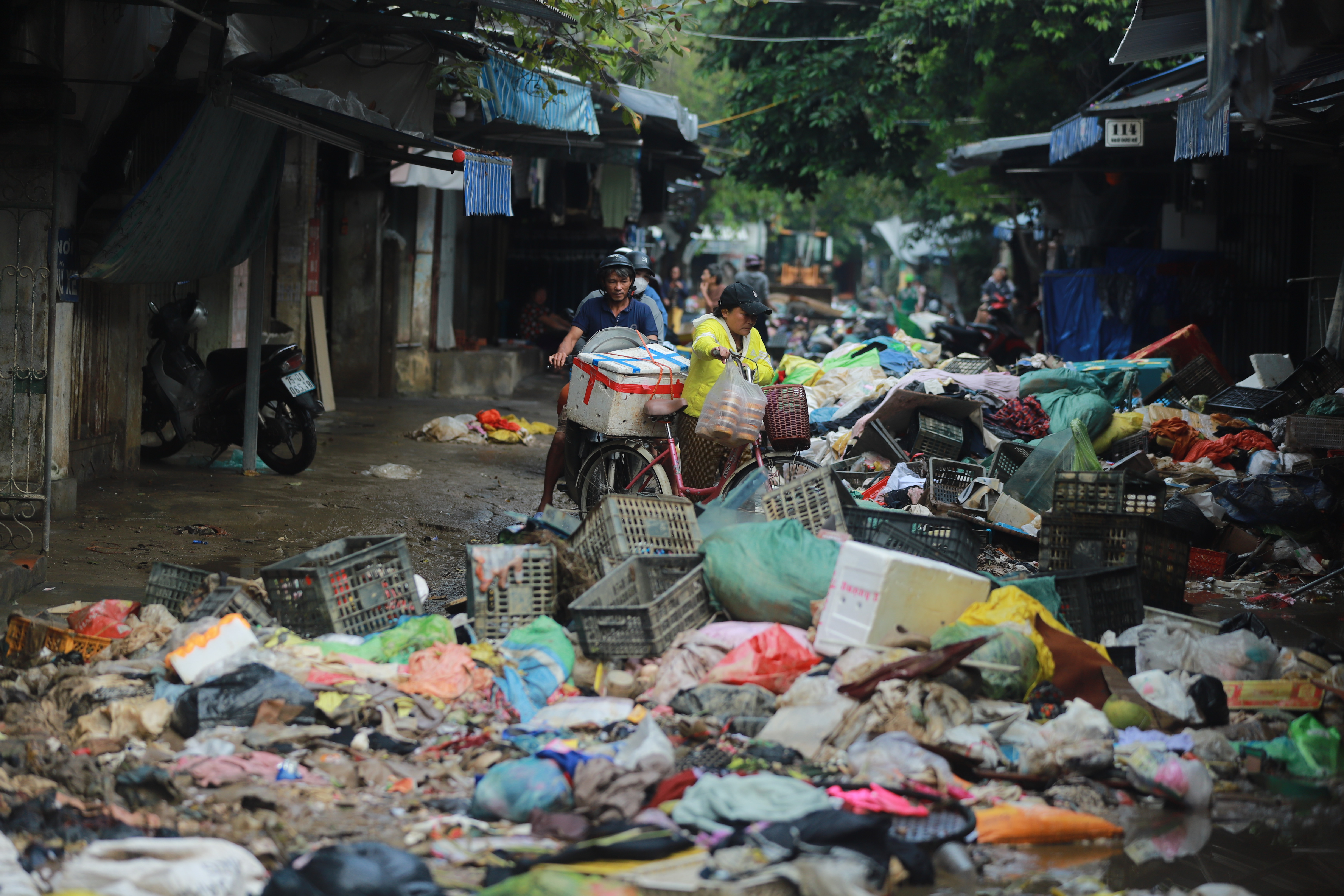 A narrow street cluttered with trash, bicycles, and people navigating through the debris.