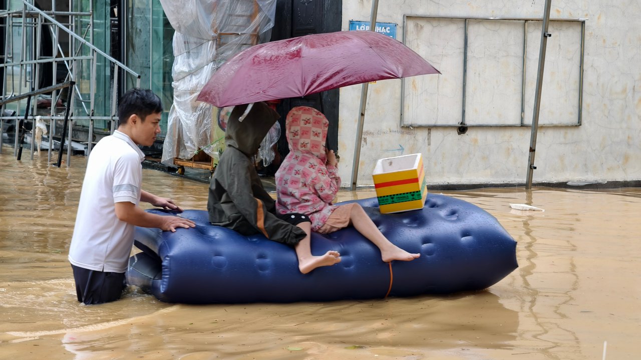 A person pulls a blue inflatable raft with two children sitting on it, under an umbrella, in a flooded area.