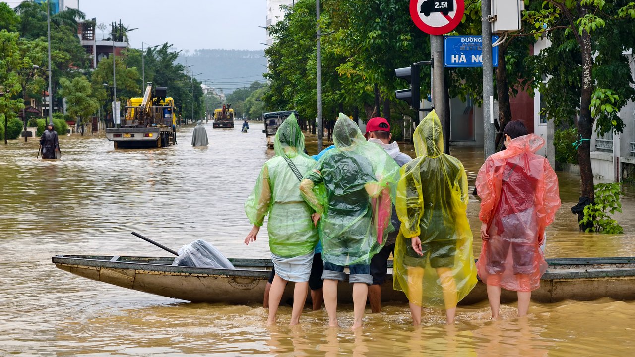 A flooded street with people walking, cars submerged, and buildings in the background under overcast skies.