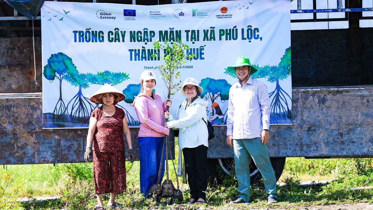 Four people stand in front of a banner promoting mangrove planting, holding a young tree in a green outdoor setting.
