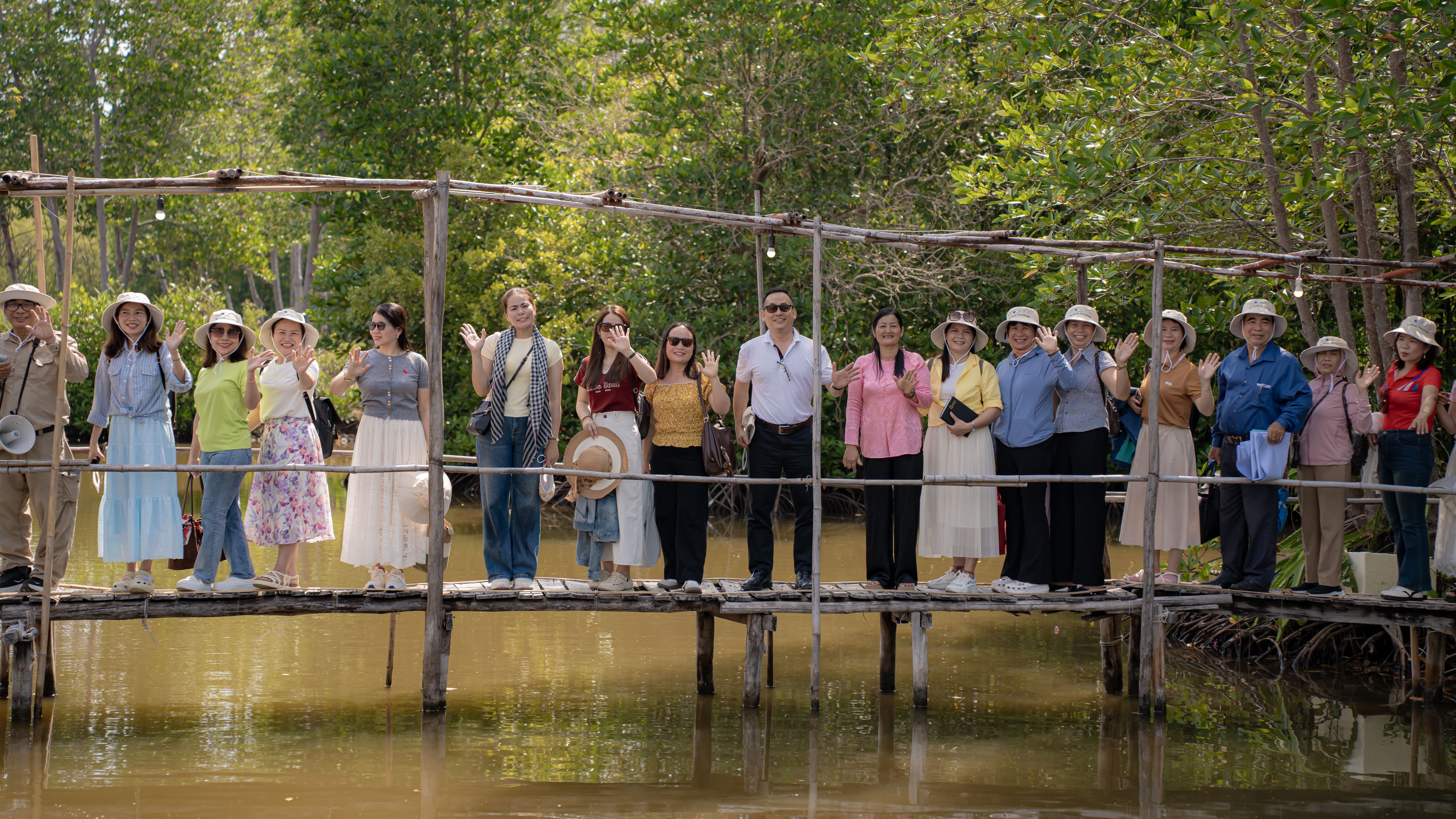 A group of people stands on a wooden bridge over water, surrounded by greenery, some waving and smiling.