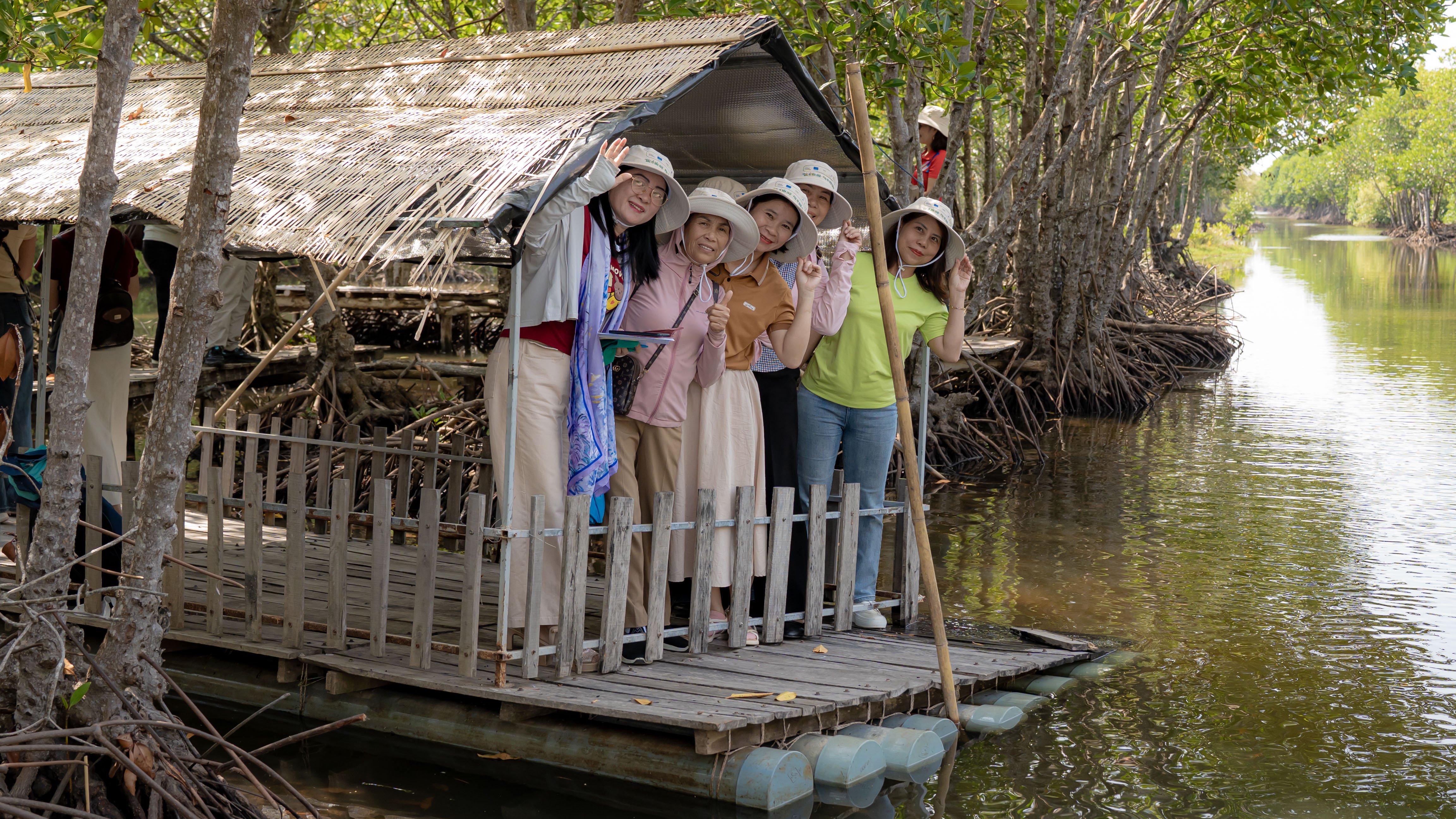 A wooden platform extends over calm water, surrounded by lush mangrove trees. Several individuals gather on the platform, wearing light-colored hats and casual clothing, engaging with each other and posing for a photo. The atmosphere is cheerful, highlighting a moment of connection with nature.
