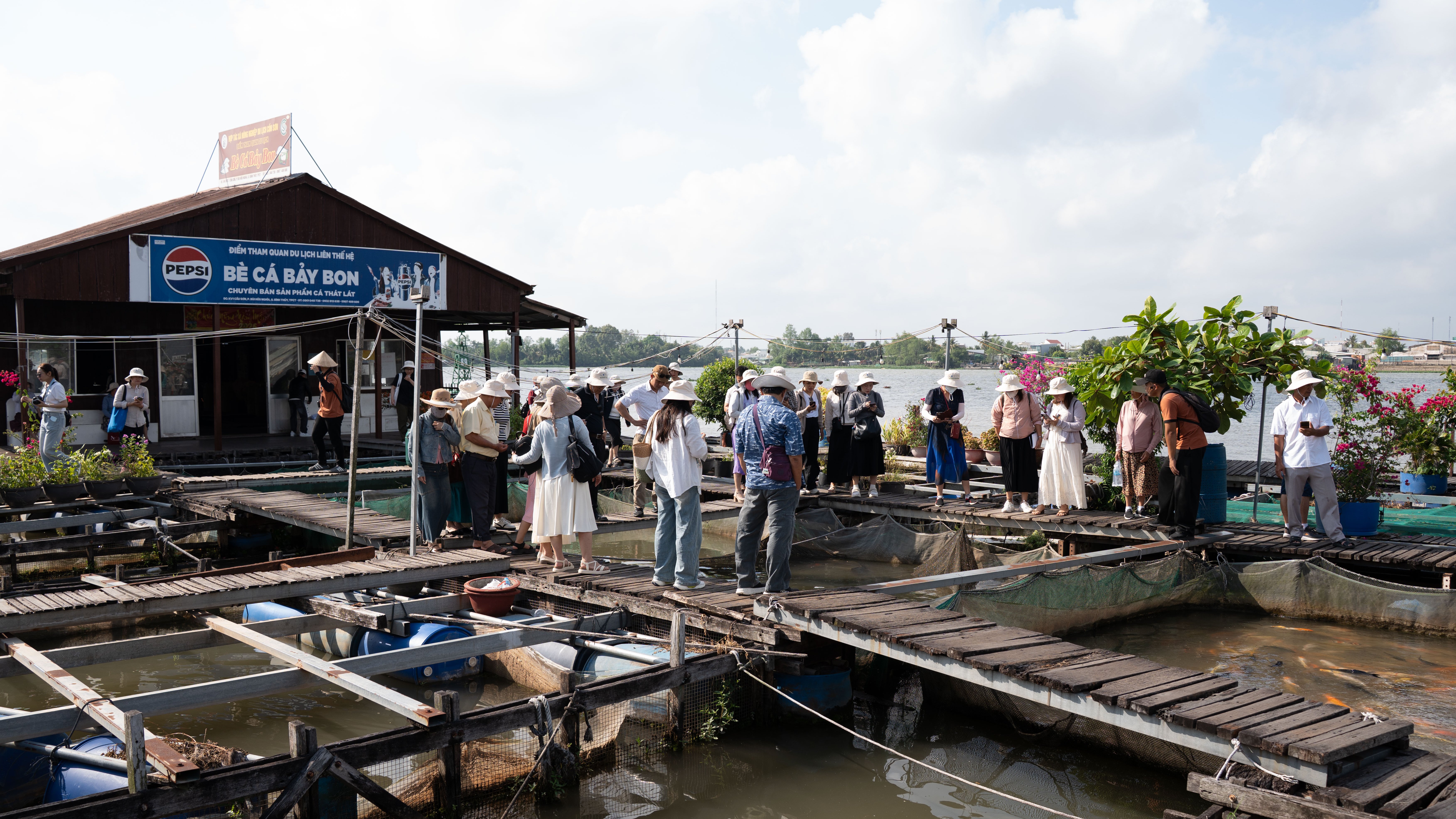 A group of people in hats gathers near a wooden structure by a waterway, with fish ponds and greenery visible.