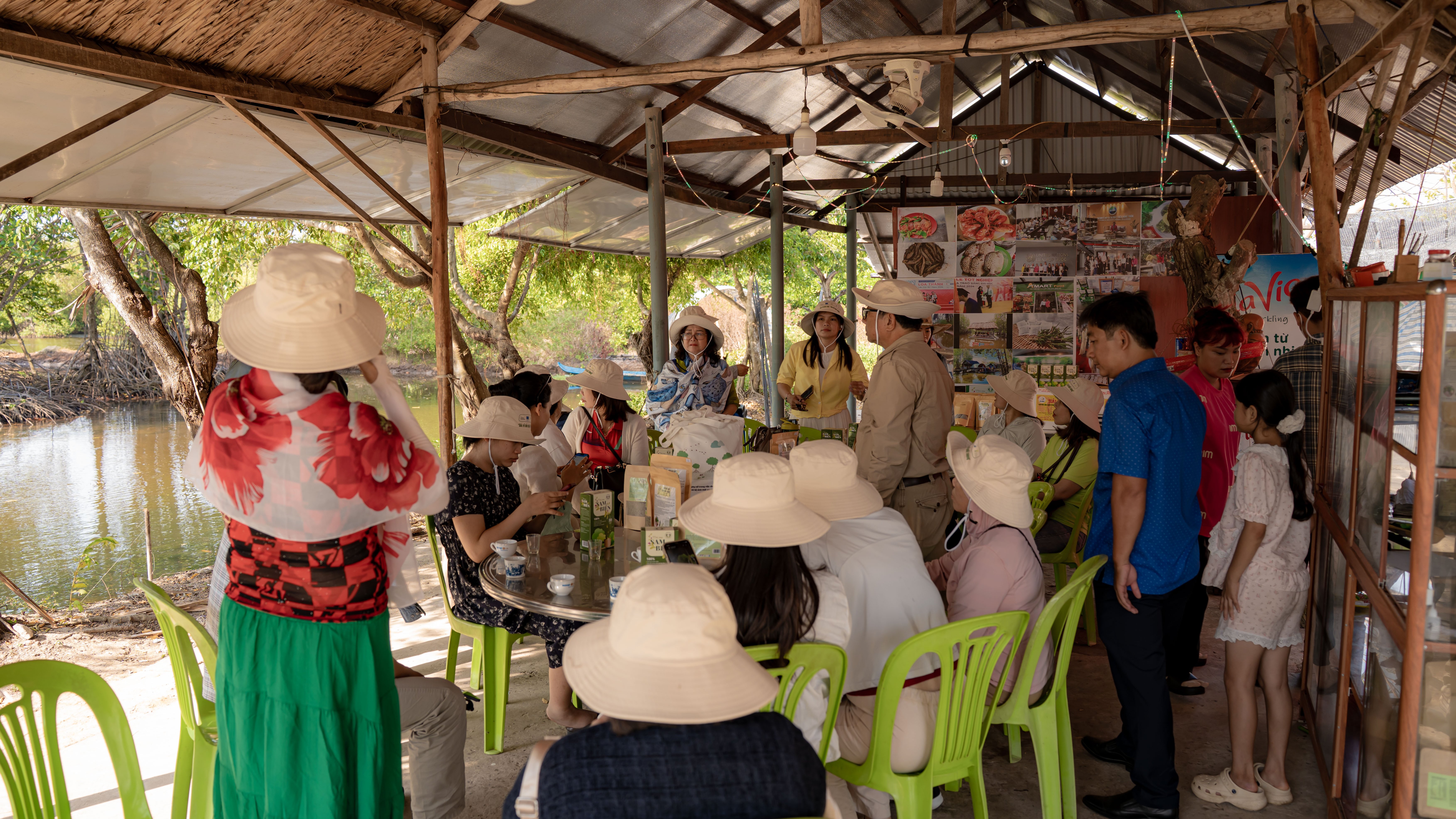 A group of people in a shaded area by a river, seated at tables, wearing hats and engaged in conversation.